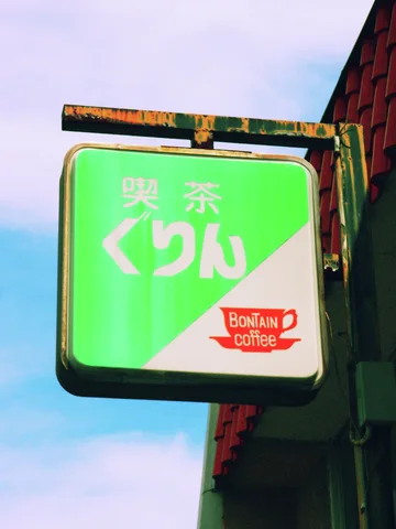 A green and cream coffee sign projects from a rusty bracket against pale blue sky.