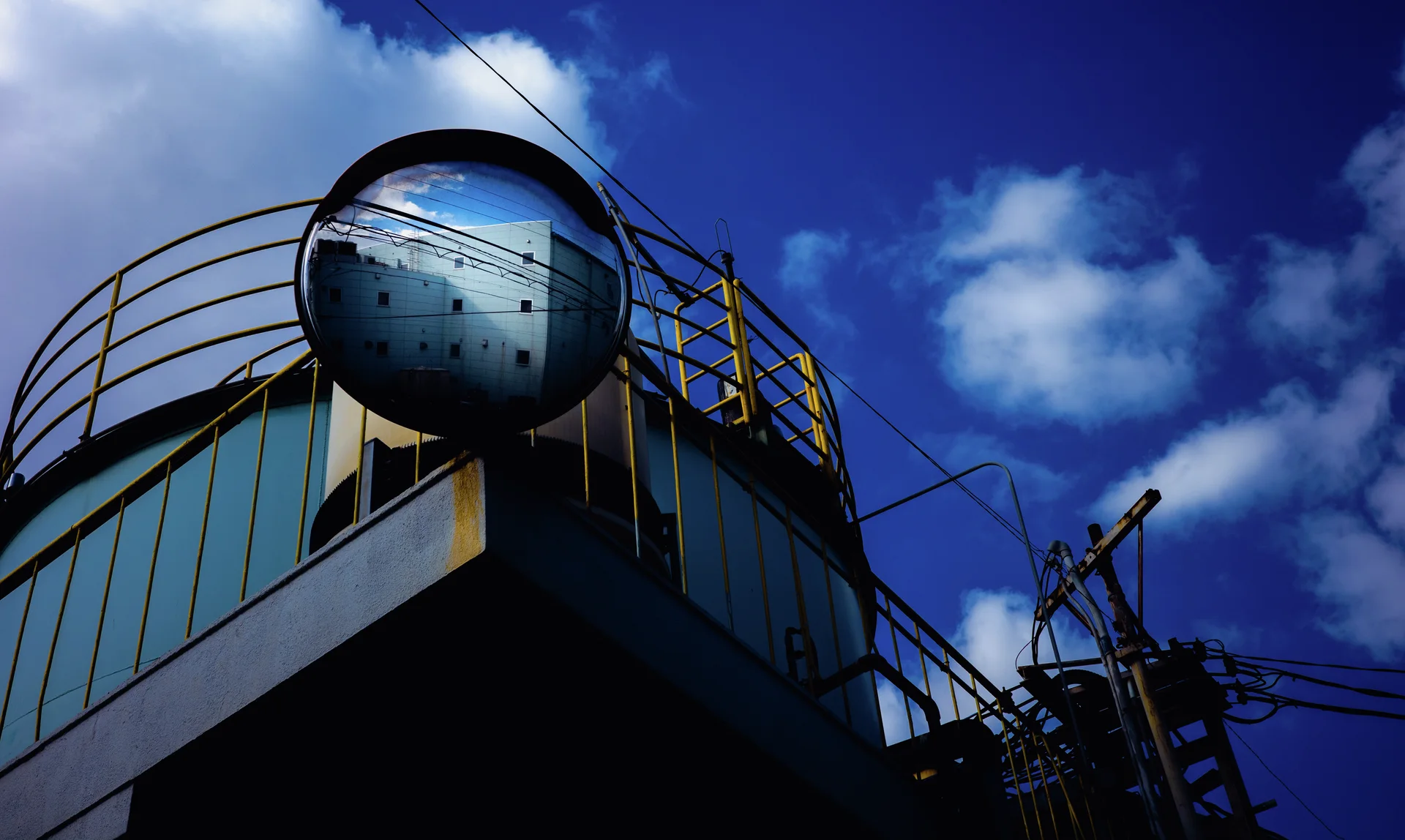 A round convex mirror reflects a pale building above yellow rooftop railings beneath blue sky and clouds.