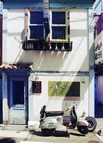 A pale scooter is parked outside a narrow white building with a blue door and upper windows.
