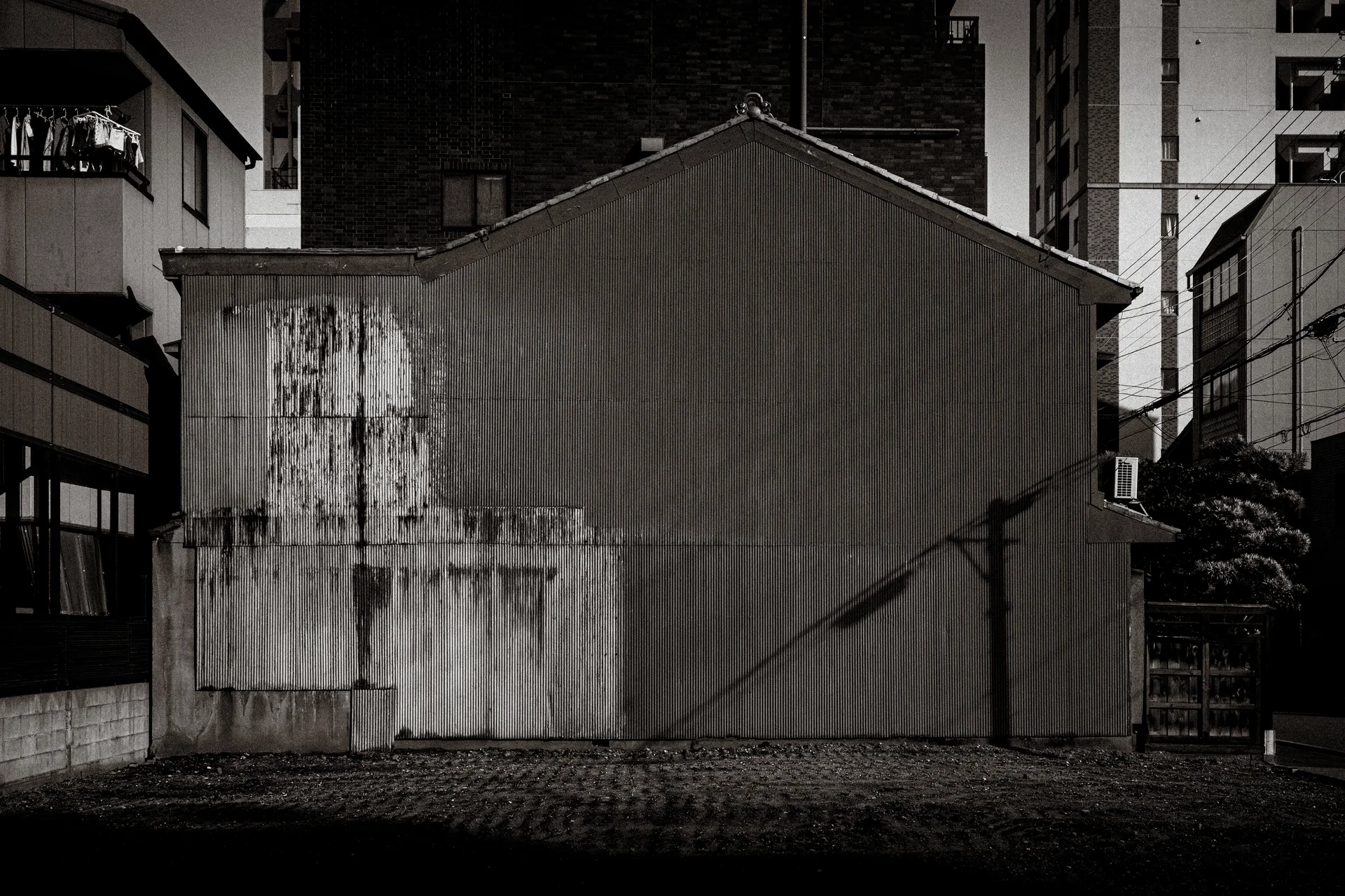 A stained blank gabled wall stands between taller buildings above patterned paving in black and white.