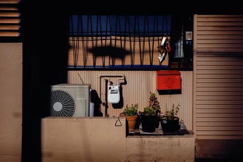 A red mailbox and utility meter sit below a barred window beside potted plants and an air conditioner.