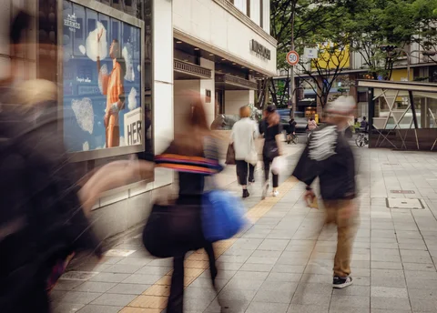Blurred pedestrians cross a broad sidewalk beside shop windows, trees, and a bright yellow tactile paving strip.