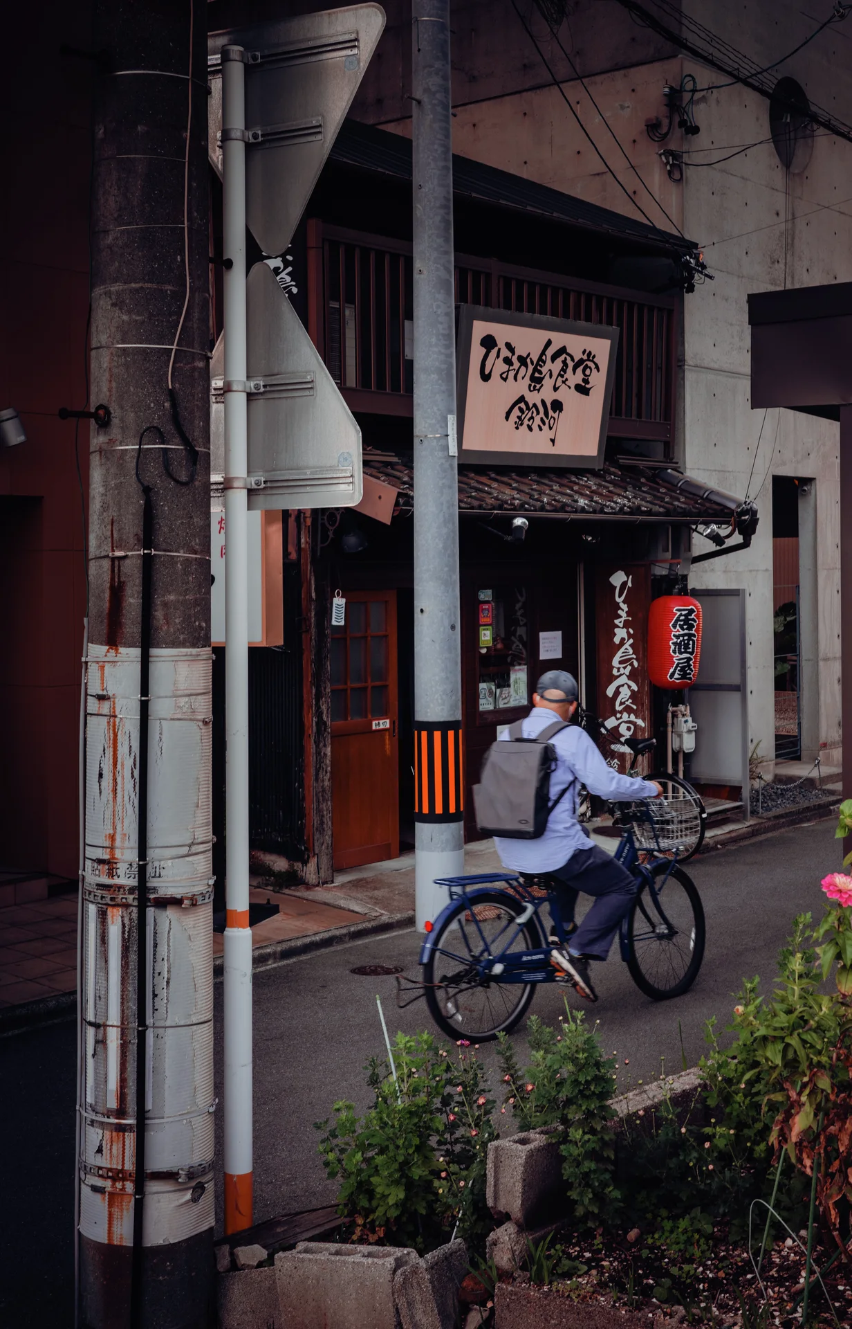 A cyclist passes a shadowed storefront with a red lantern beside a utility pole.