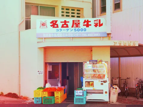 Colorful crates and a vending machine stand outside a pale storefront beneath a red-lettered sign.