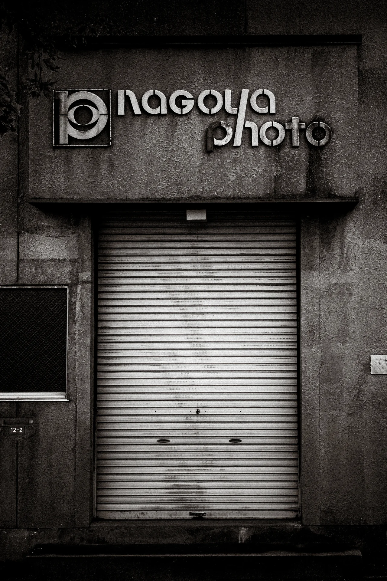 Raised Nagoya photo lettering sits above a closed shutter on a weathered monochrome facade.
