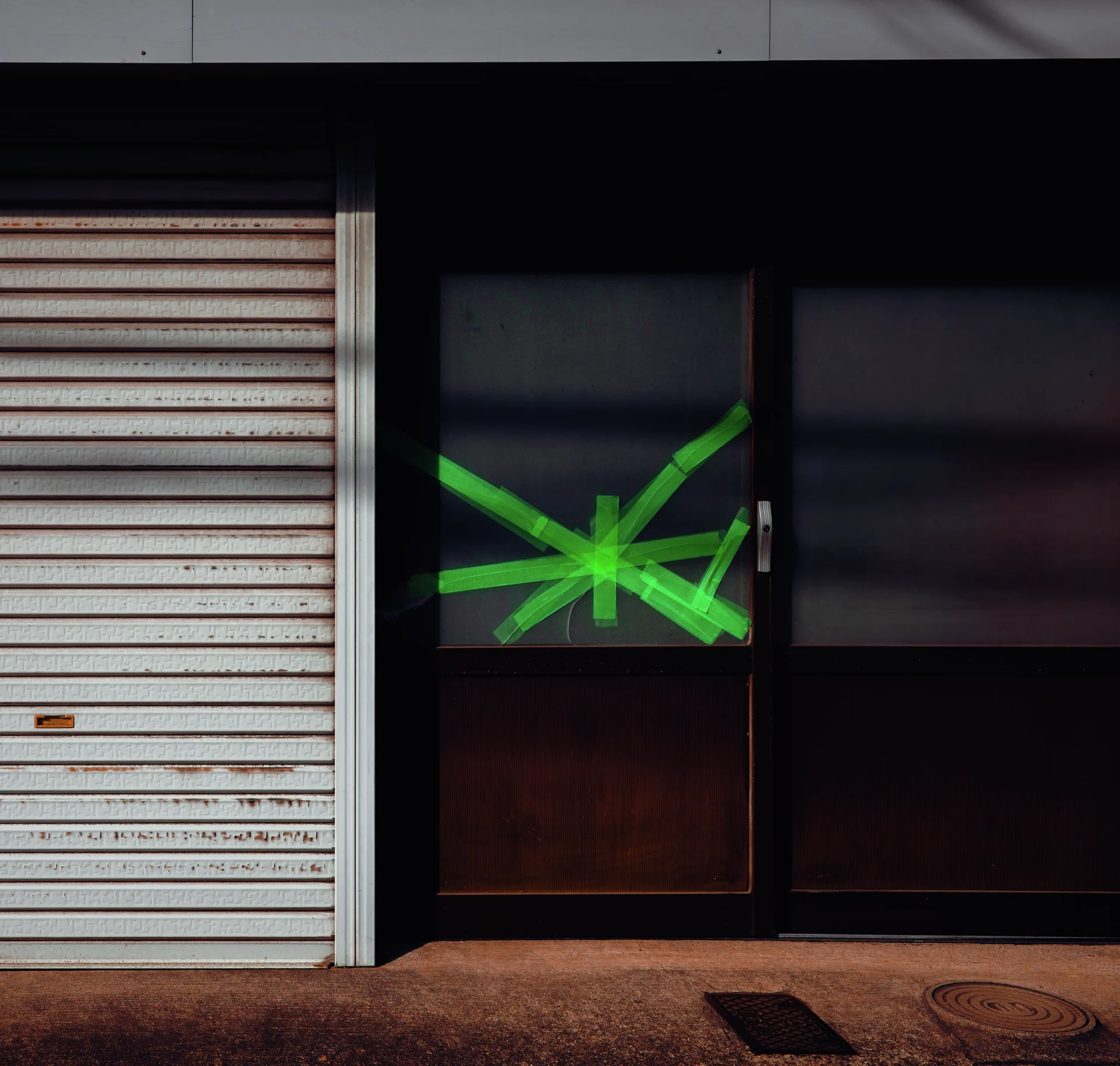 Bright green tape forms a starburst across dark glass doors beside a closed pale shutter.