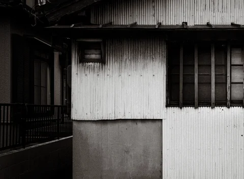 A weathered corrugated wall has small dark windows beside a shadowed narrow passage in black and white.