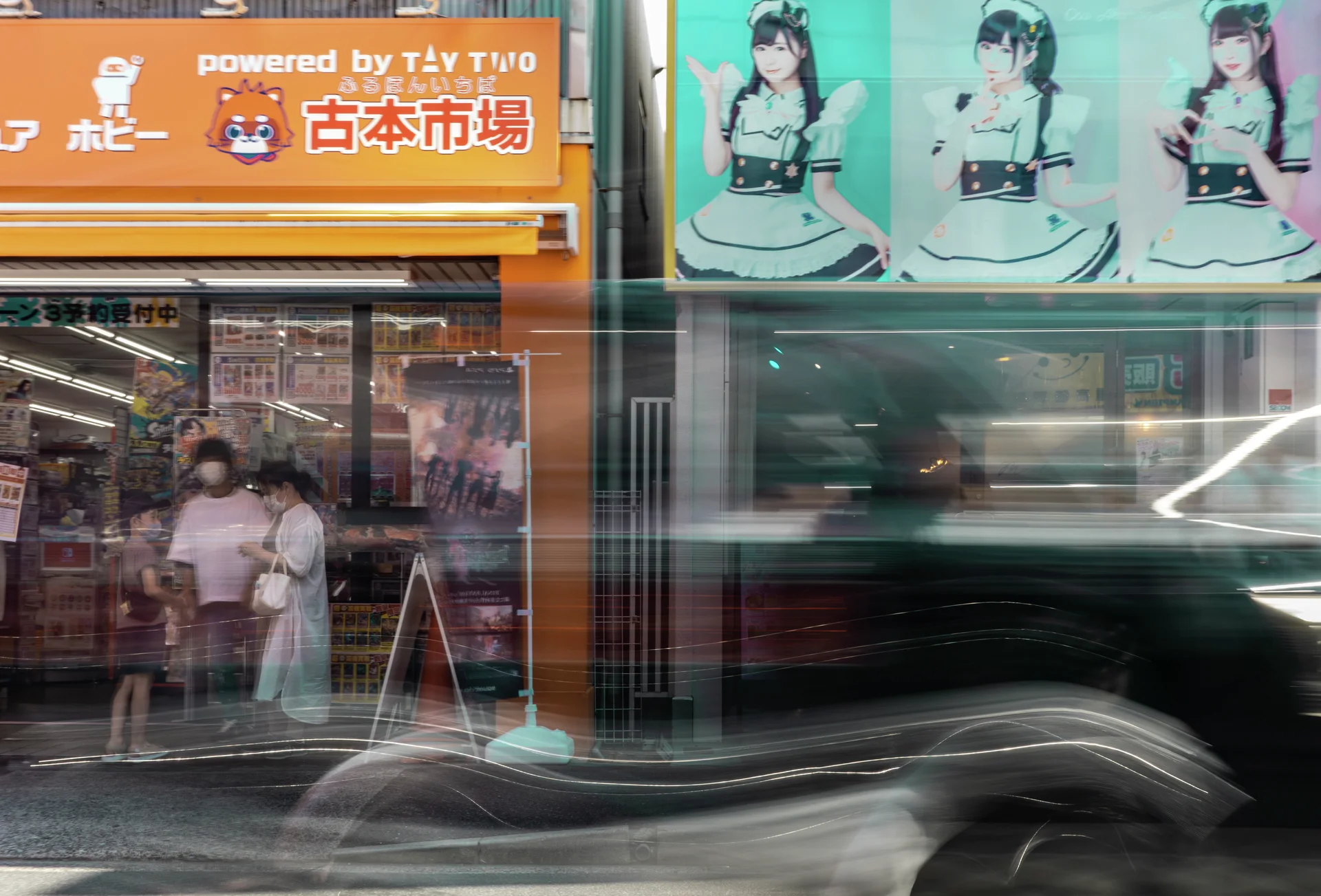 A blurred dark vehicle passes storefront windows beneath an orange sign and illustrated character posters.