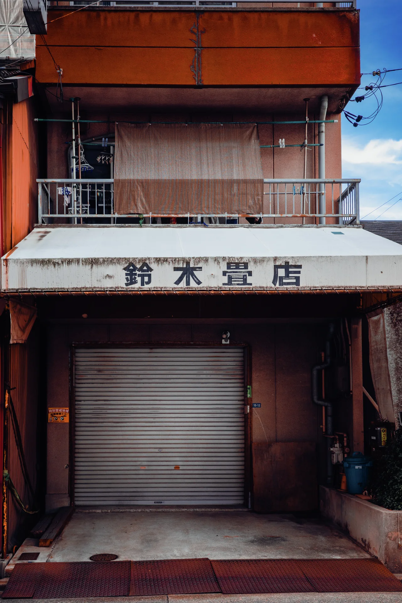 A closed metal shutter sits below a white Japanese sign and an orange upper balcony.