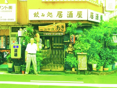 A person stands outside a restaurant entrance surrounded by signs, lanterns, and dense potted greenery.