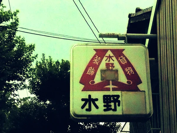 A weathered cream sign displays a red geometric emblem and dark Japanese lettering beside leafy trees.