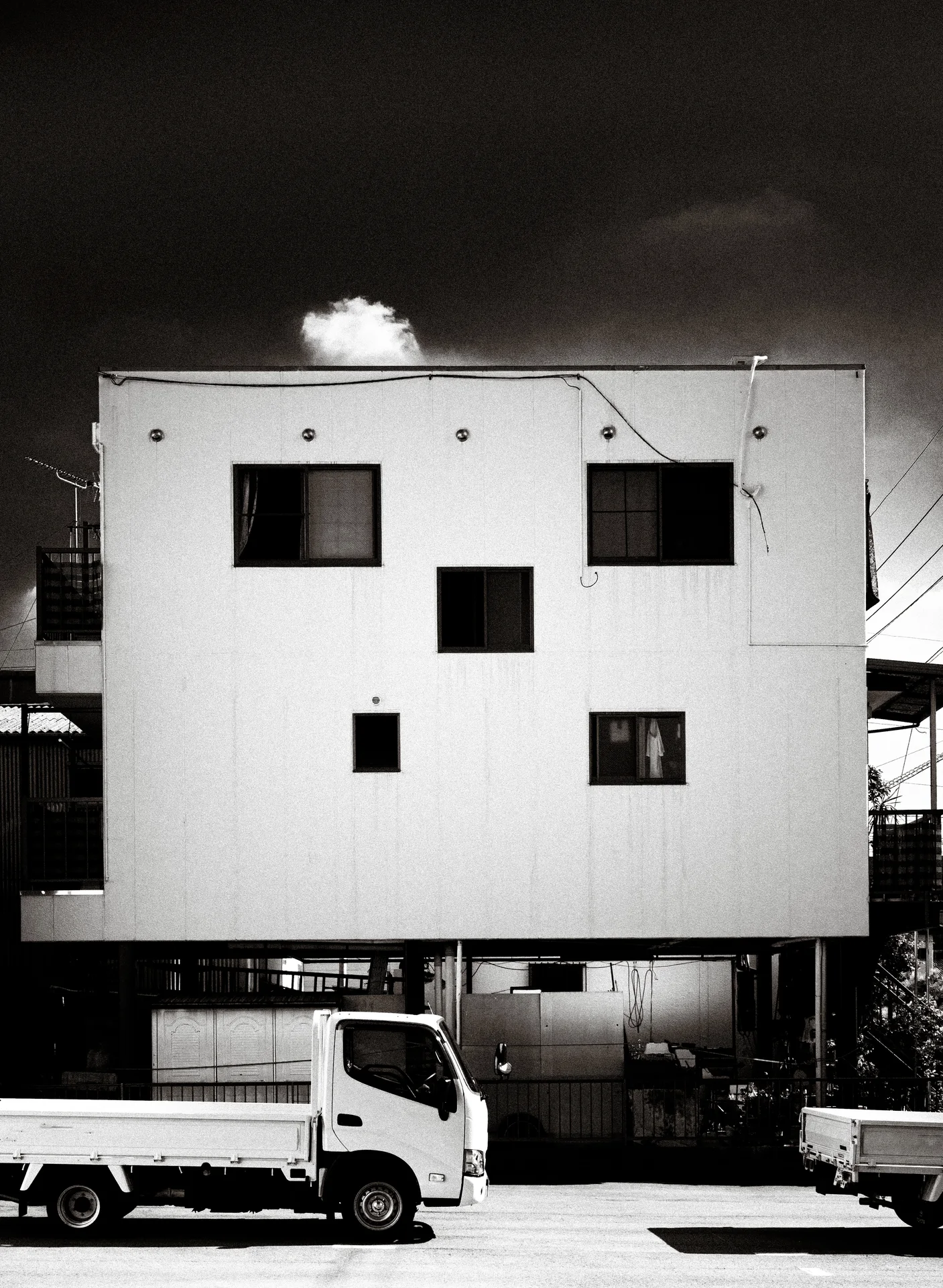 Small dark windows punctuate a pale square building above a parked truck in black and white.