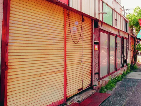 Closed beige shutters and dark windows line a weathered storefront with red trim and pavement weeds.