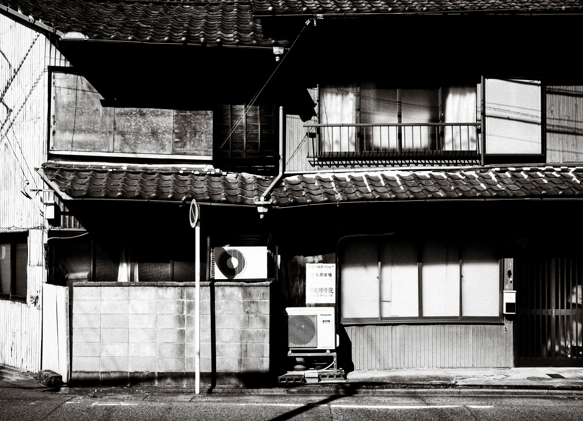 A weathered two-story house has tiled eaves, closed windows, and stacked air conditioners in black and white.
