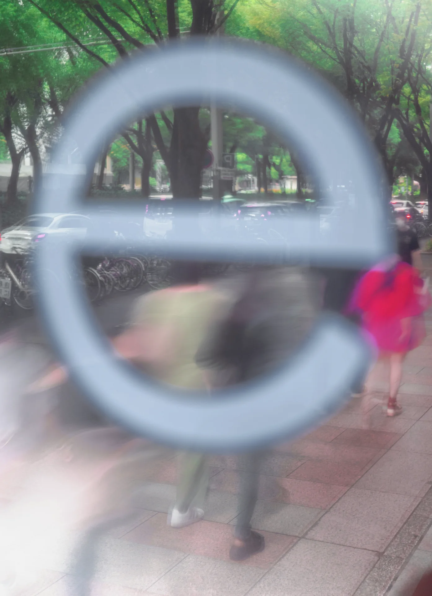 A large pale letter on glass overlays blurred pedestrians and leafy trees beyond the window.