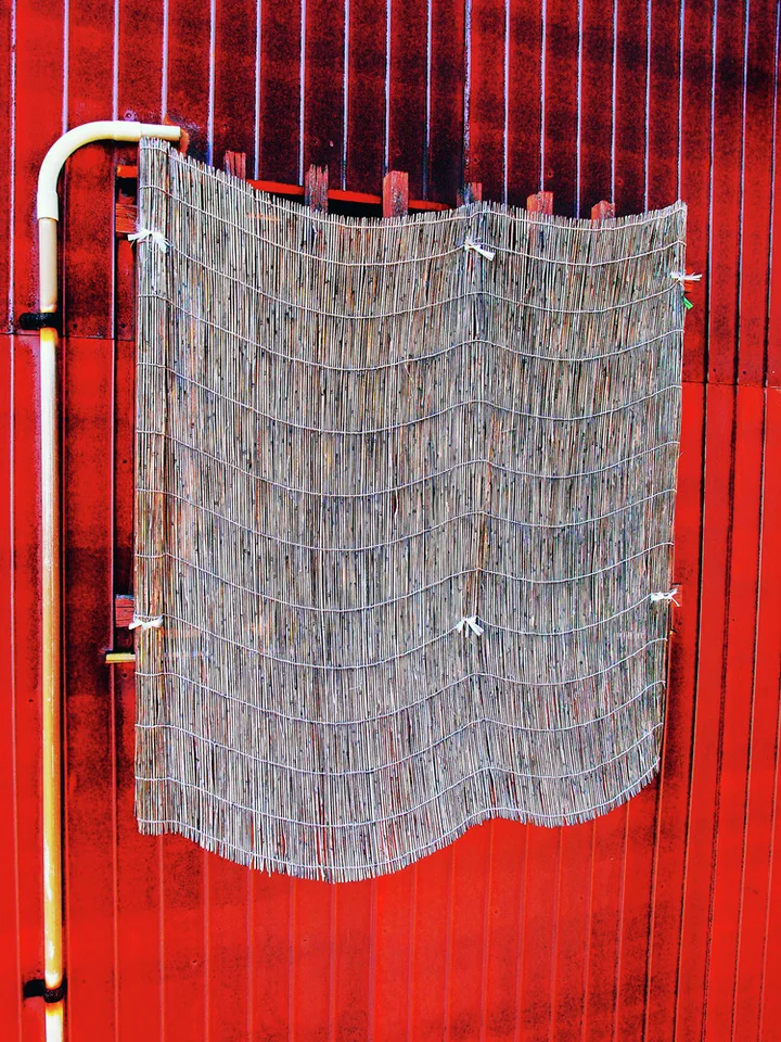 A curved gray woven screen hangs from a pale frame against bright red corrugated siding.