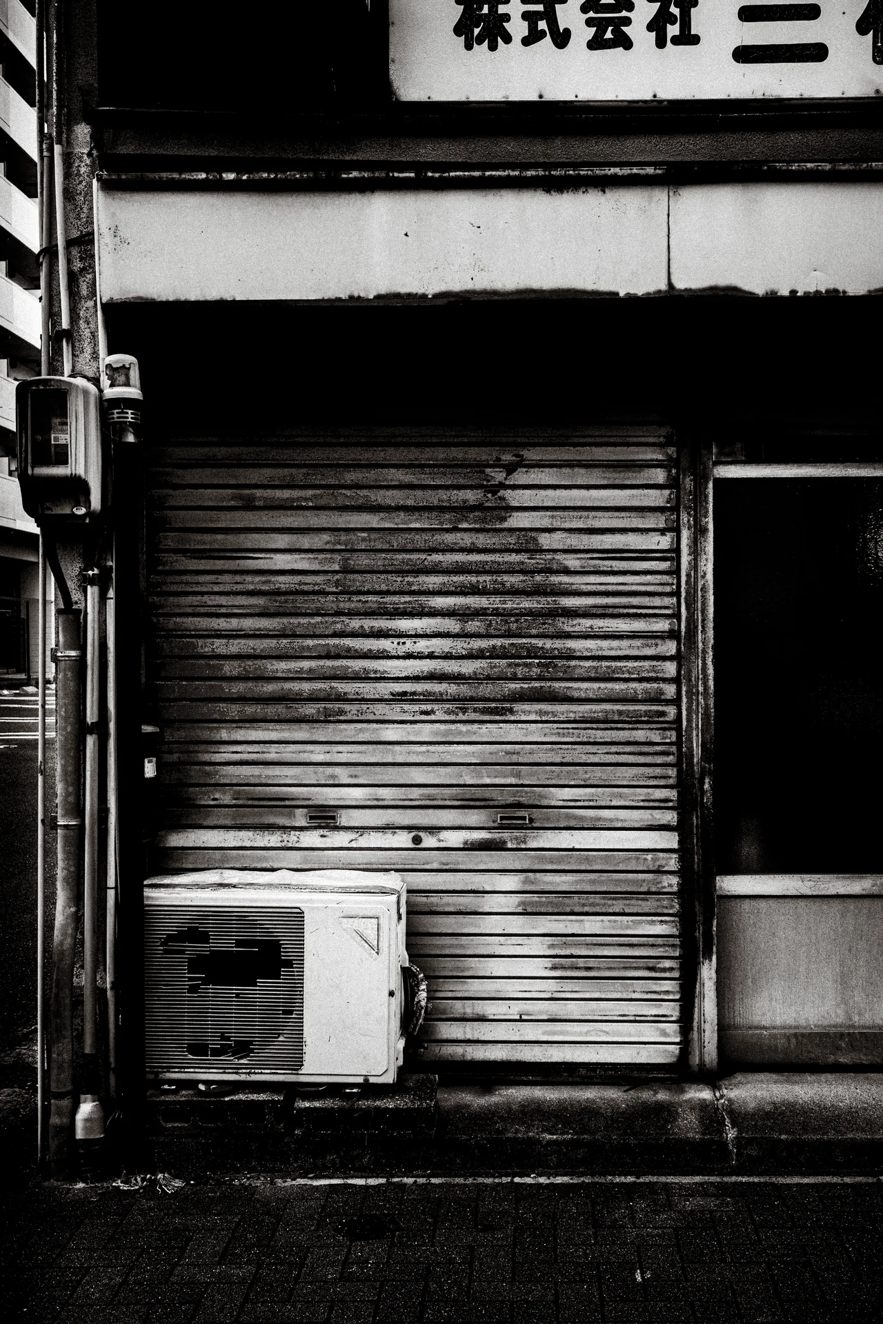 A rust-marked closed shutter stands behind an outdoor air conditioner beneath a shop sign in black and white.