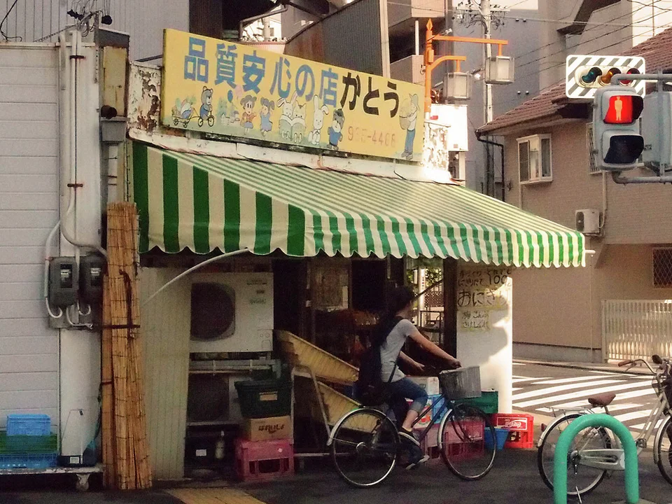 A person on a bicycle pauses outside a small shop beneath a green striped awning and faded yellow sign.