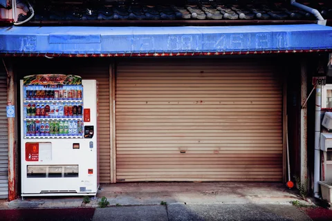 A white drink vending machine stands beside closed beige shutters beneath a faded blue awning.