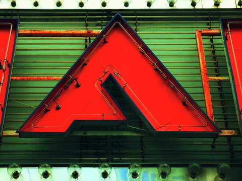 A large red triangular letter hangs against green corrugated siding between rows of round bulbs.