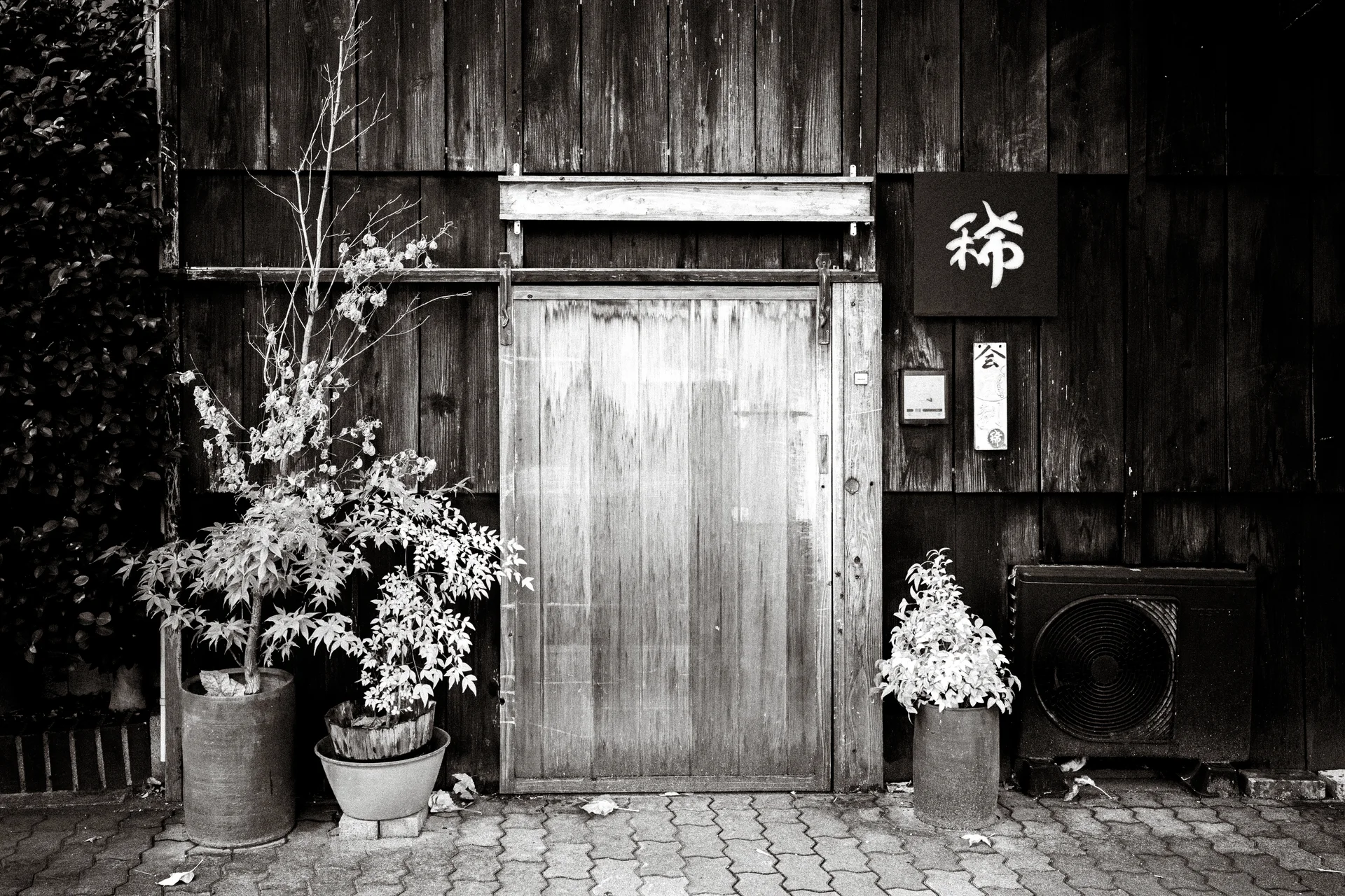Potted plants flank a pale wooden door set within a dark weathered timber facade.