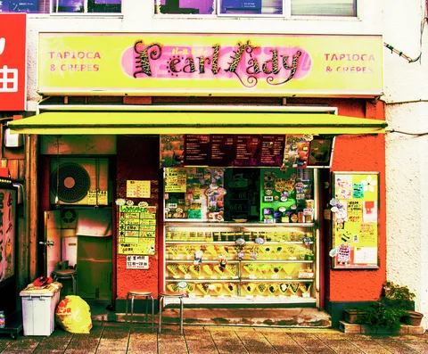 A small orange shopfront displays products in a glass case beneath a yellow awning and decorative sign.