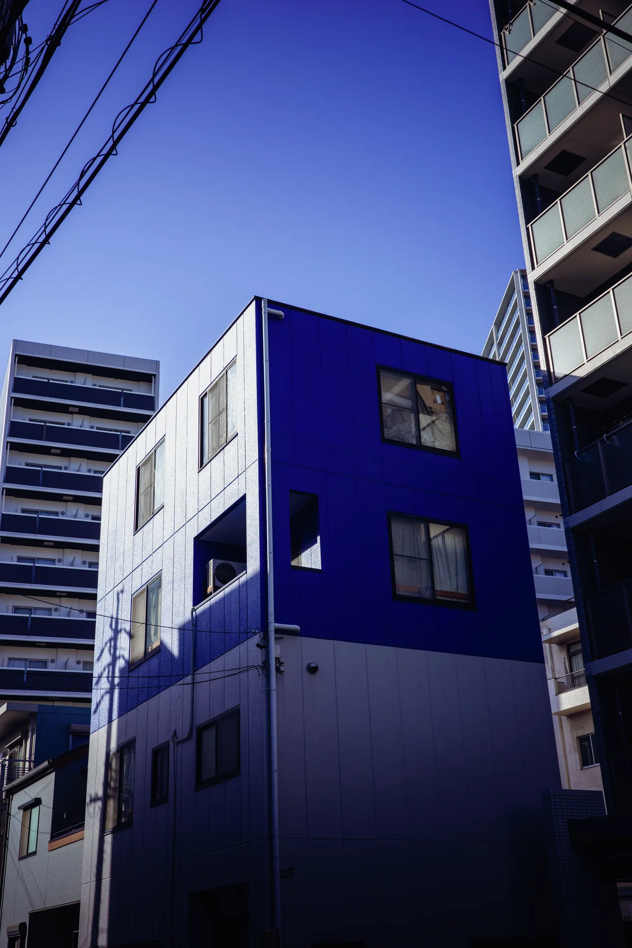 A narrow blue and pale building rises between taller apartment blocks beneath overhead cables.