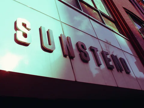 Raised SUNSTENO lettering casts shadows across pale facade panels beneath upper windows.