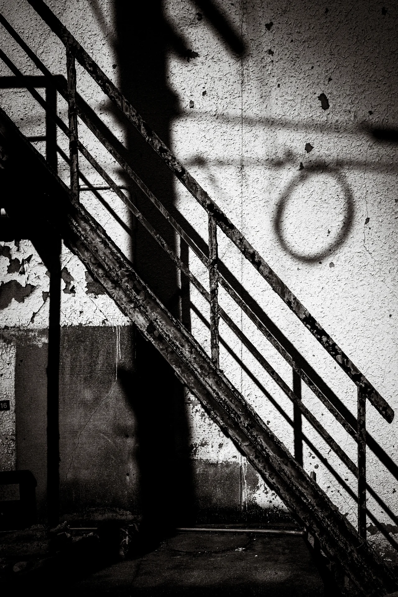 A diagonal metal stair railing casts angular shadows across a wall marked with a circle in black and white.