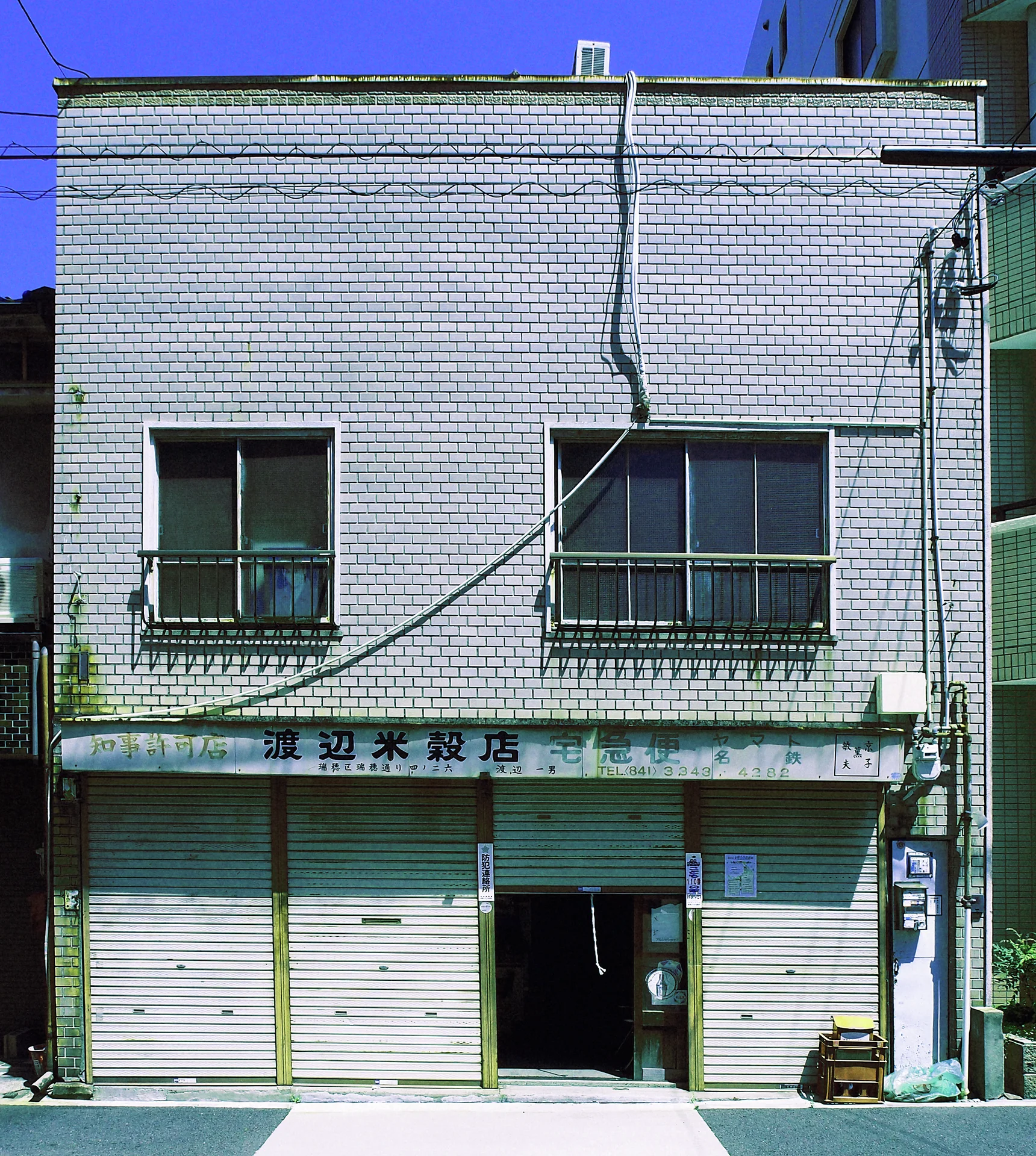 A pale tiled building has two upper windows above three closed shutters and a narrow open entrance.