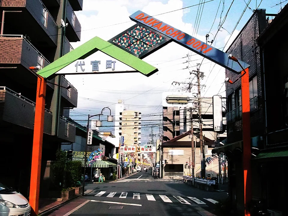 Orange posts support angular green and blue street gateways above a narrow road crossed by utility wires.