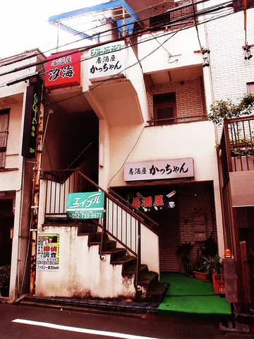 An exterior staircase rises above a narrow restaurant entrance with green matting and small signs.