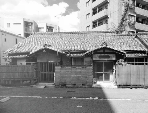 A low tiled-roof house and block wall stand before taller apartment buildings in black and white.