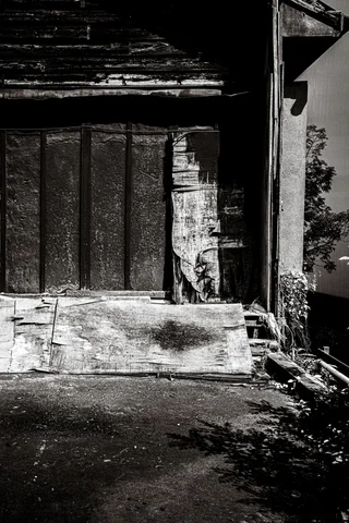 Sunlight strikes rough wooden boards and crumbling wall edges above uneven ground in black and white.