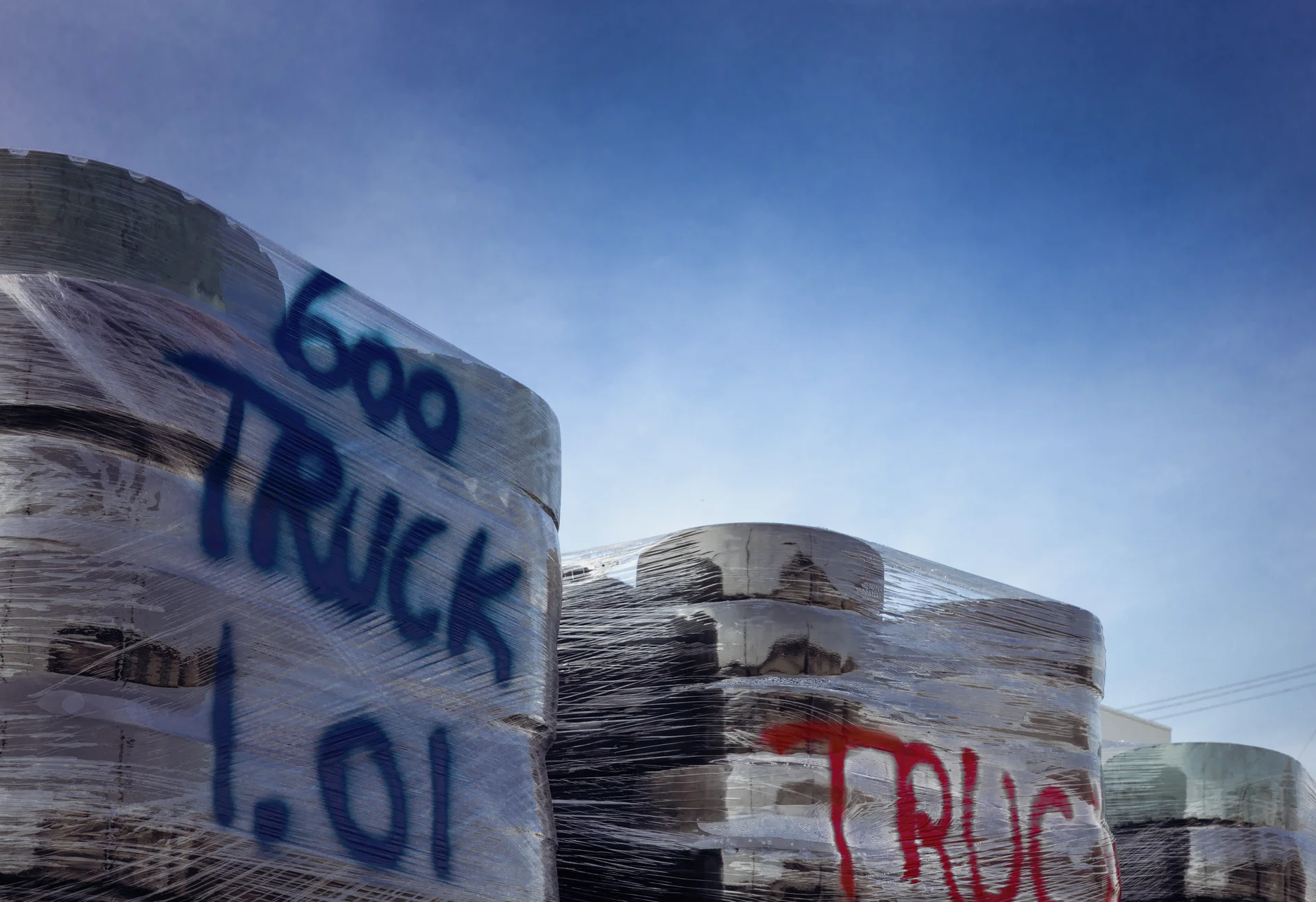 Stacks of tires wrapped in clear plastic display large blue and red markings beneath pale blue sky.