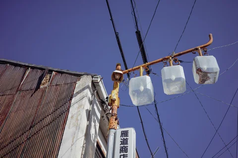 Three cloudy rectangular lamps hang from a rusty curved bracket beneath overhead wires and blue sky.