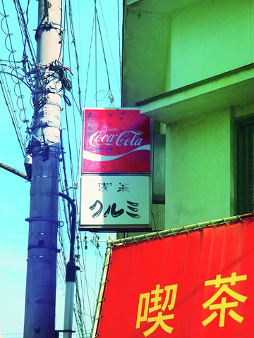 A red Coca-Cola sign projects beside a utility pole above a red awning with yellow Japanese lettering.