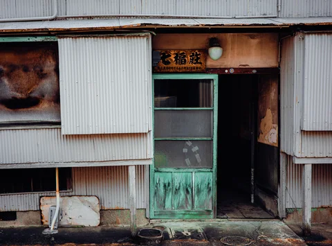 A worn turquoise door opens into darkness amid patched corrugated siding and stained walls.