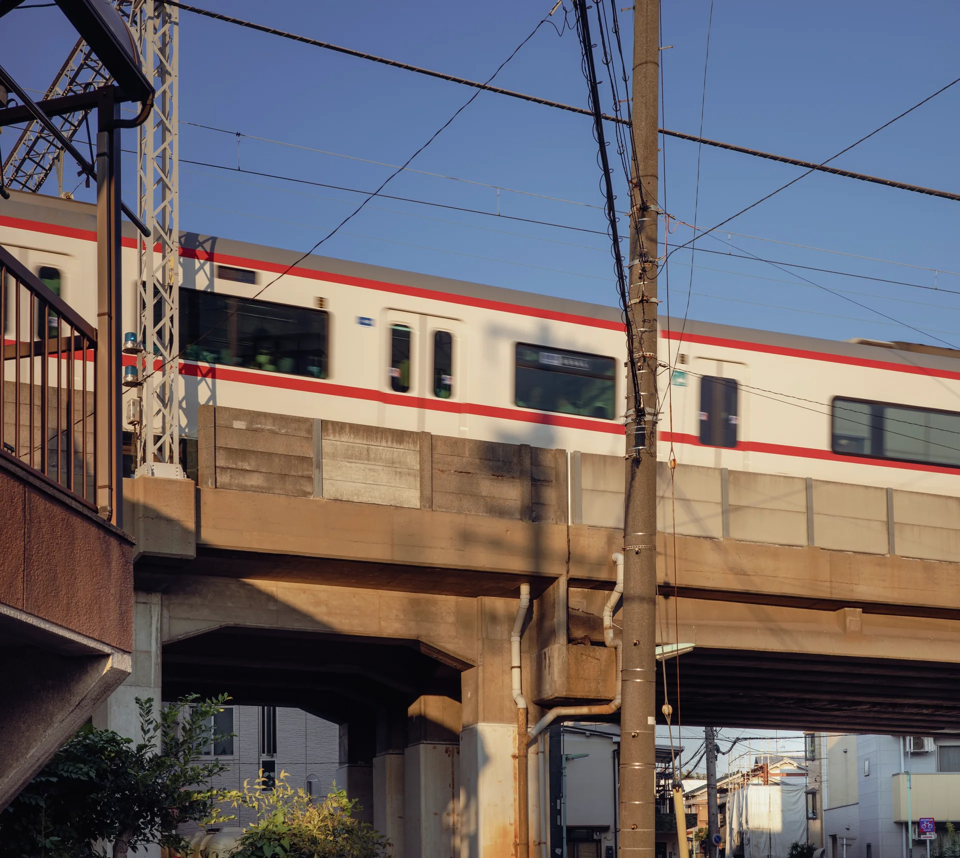 A white train with a red stripe crosses an elevated concrete track behind utility poles and wires.