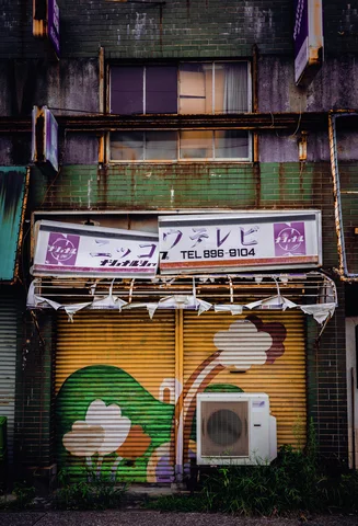 A narrow weathered building has colorful painted shutters beneath shop signage and a green balcony railing.