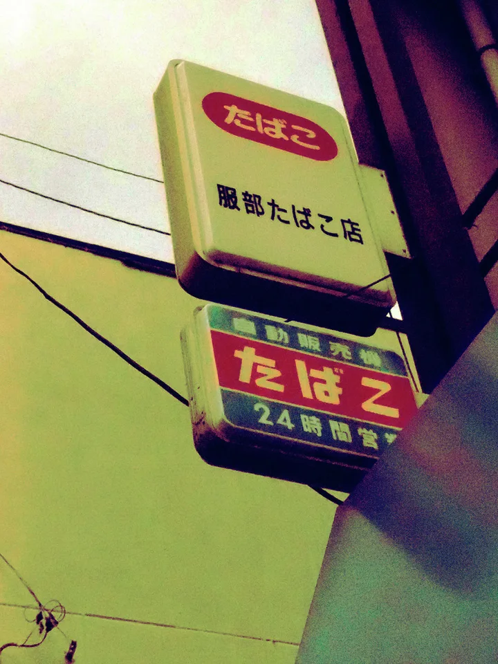 Two small cream tobacco signs project beside a dark building beneath wires and pale yellow sky.