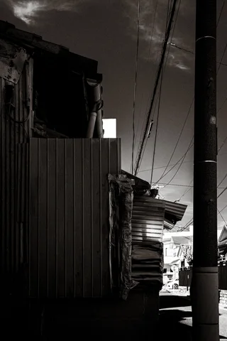 A narrow shadowed passage runs between weathered corrugated buildings and a utility pole in black and white.