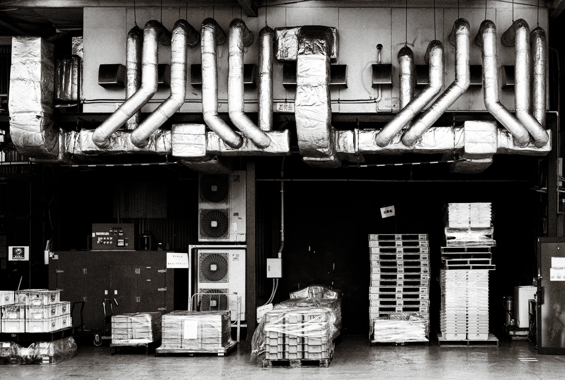 Rows of bent ventilation ducts line an industrial facade above stacked pallets and containers in black and white.