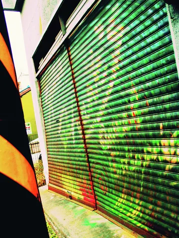 Mottled green paint and rust mark closed metal shutters beside a narrow passage.