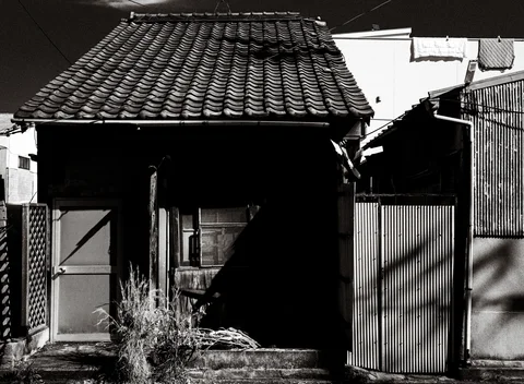 A small tiled-roof house has a deeply shadowed entrance and overgrown steps in monochrome.