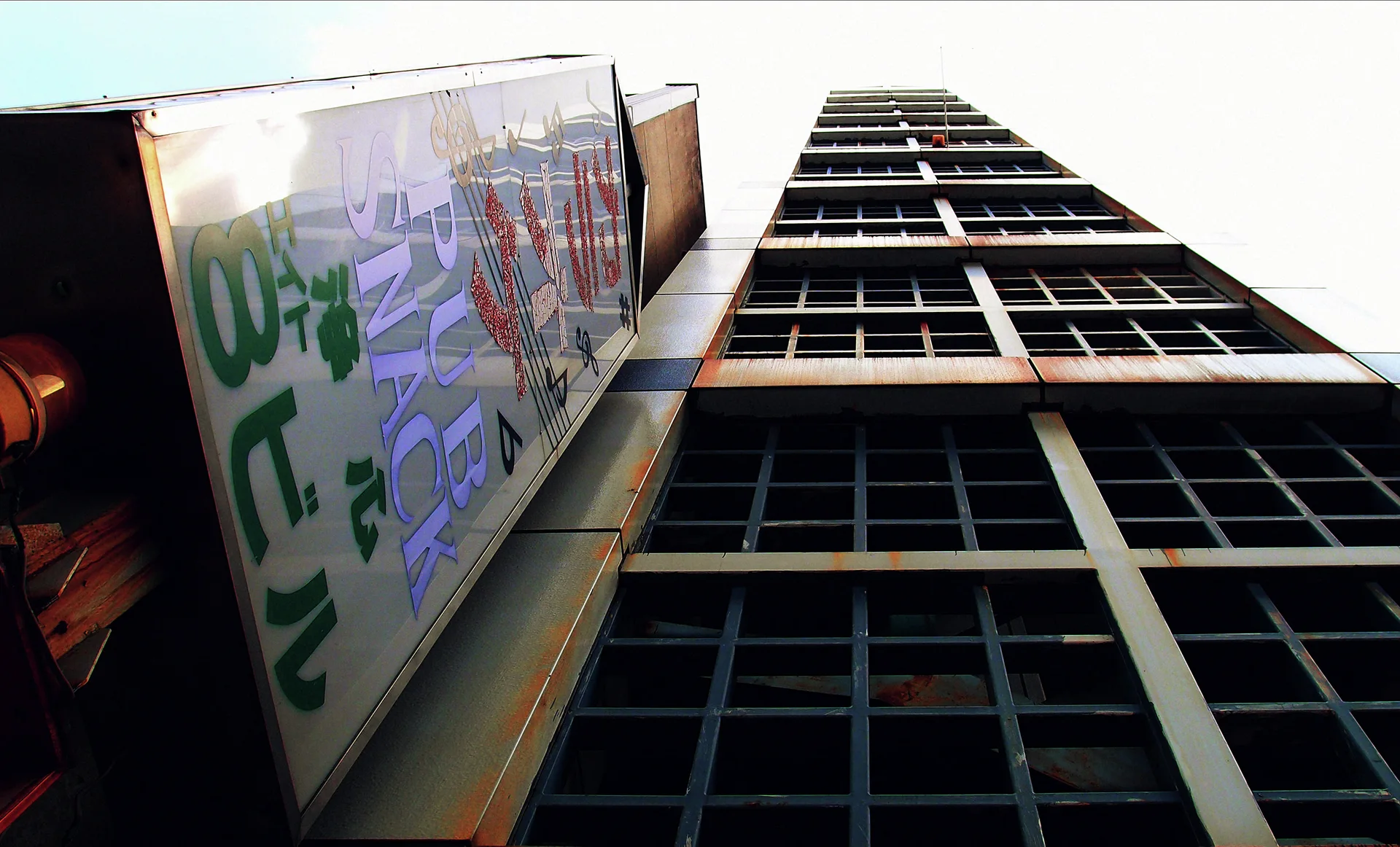 A steep upward view follows a gridded high-rise facade beside a large projecting sign.