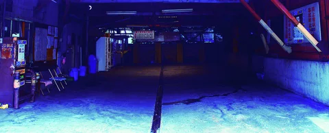 A dark covered passage contains empty chairs and pipes above blue pavement divided by a narrow drain.