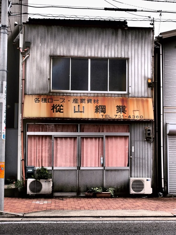 A narrow corrugated shop has a faded tan sign above pink curtains and two ground-level air conditioners.