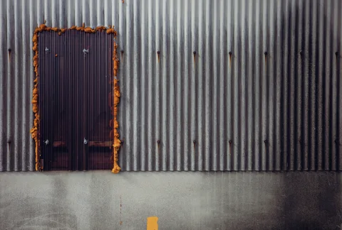 Orange rust outlines a small dark metal door set into a gray corrugated wall.