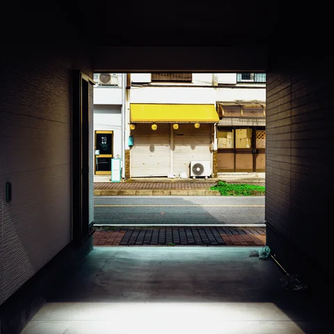 A shadowed passage frames a sunlit street and a closed storefront beneath a bright yellow awning.